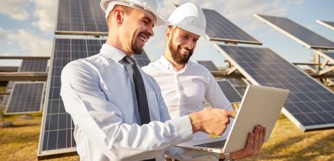 Engineer using laptop near photovoltaic panels producing renewable solar energy Positive male technicians in formal wear and hardhats discussing configuration of solar panels while standing close and using netbook together